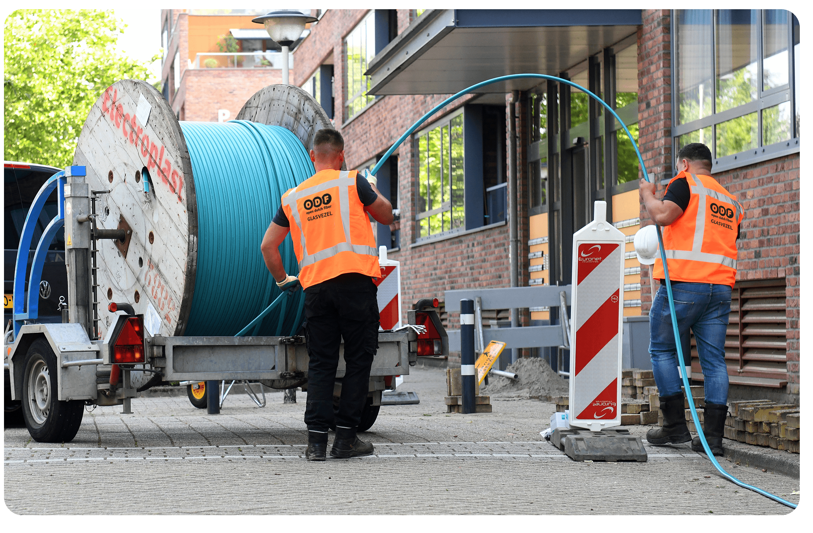 Twee werknemers in oranje veiligheidsvesten trekken een grote blauwe kabel van een haspel op een aanhangwagen naar een gebouw aan de kant van de weg.