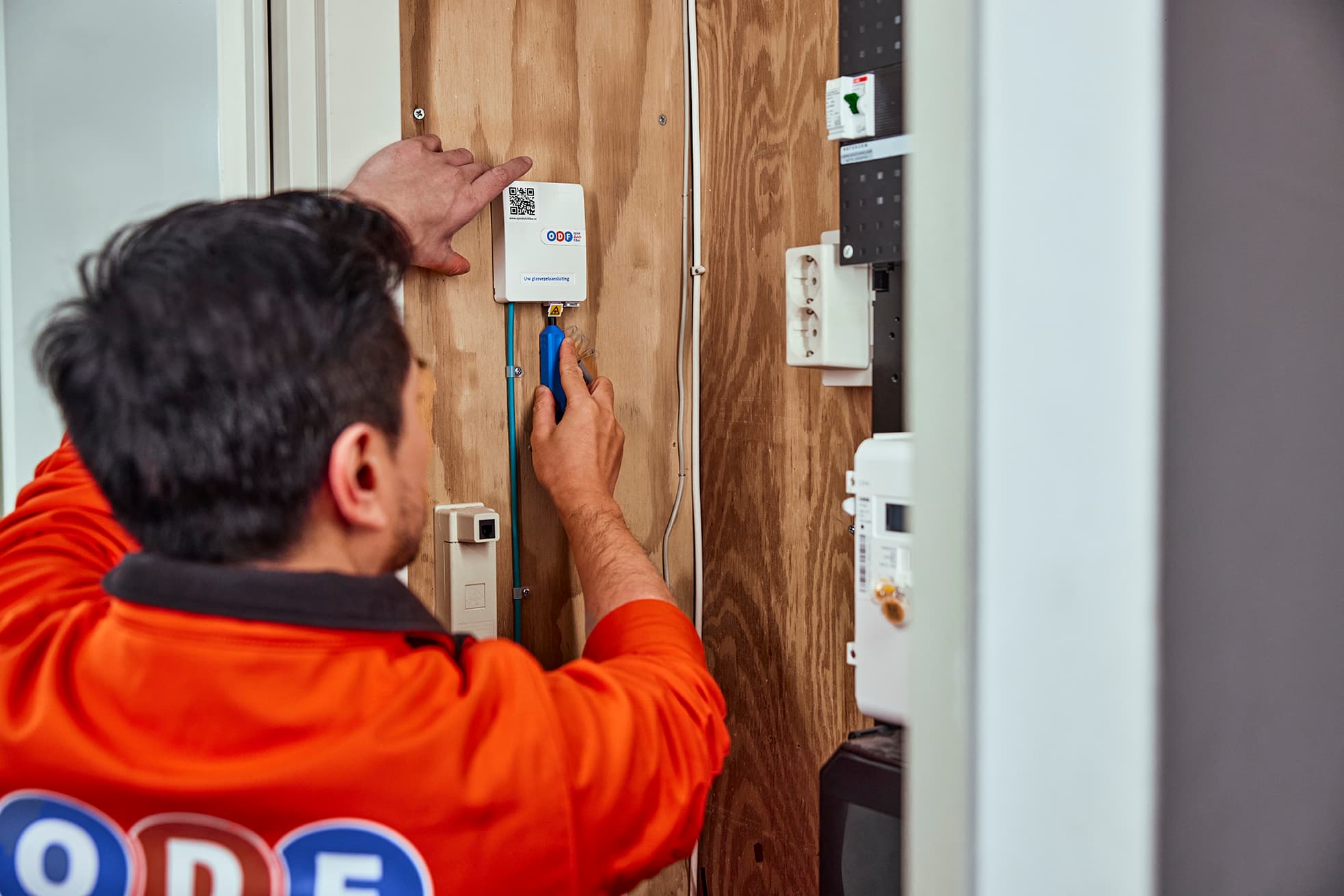 Montering van een witte communicatie- of internetbox (met QR-code) door een technicus in oranje T-shirt met logo van ODF, bezig met glasvezelinstallatie in een meterkast of technische ruimte.